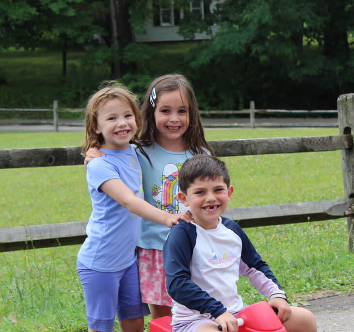 Three young campers in front of a fence, two girls standing behind a boy on a tricycle